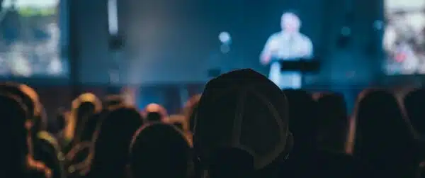 An audience looking towards a stage where a man is talking. Everything is blurry except the person closest to the camera.