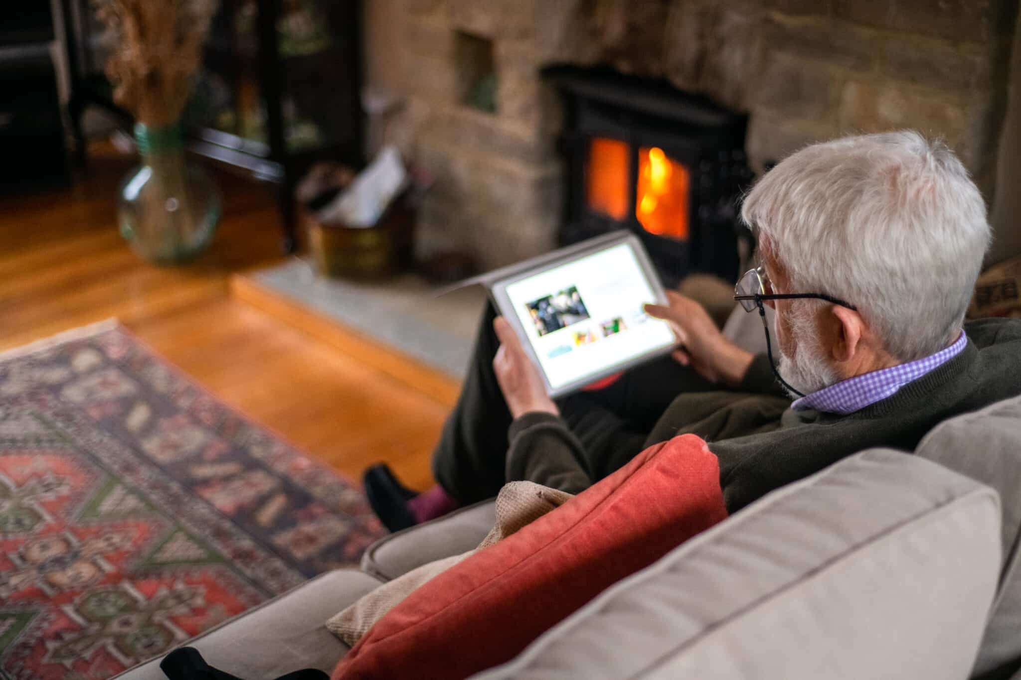 Old man surfing a website with an tablet is sitting in a beige sofa with a cosy fireplace. The man has gray hair and glasses.