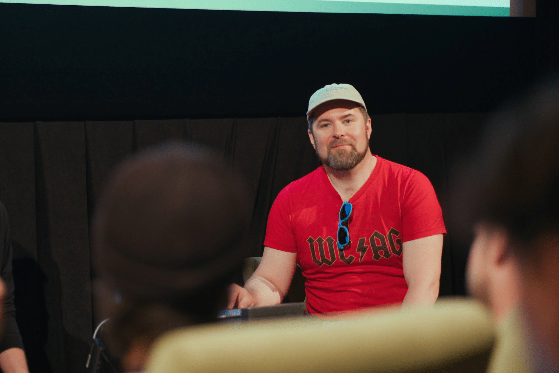 Erik sitting in front of an audience, wearing a beige cap and red T-shirt with the text ‘WCAG’ in an AC/DC-style font