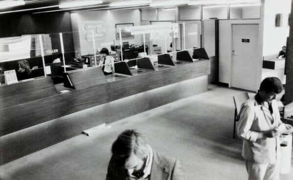 Black-and-white photo of a bank interior with teller counters and customers being served.
