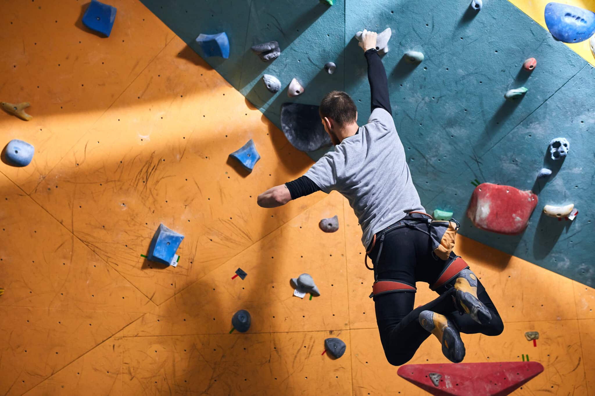 A man rock climbing indoors, holding his one hand on a grip while hanging freely. The wall is painted in yellow and turquoise with blue and red grips in different shapes. 